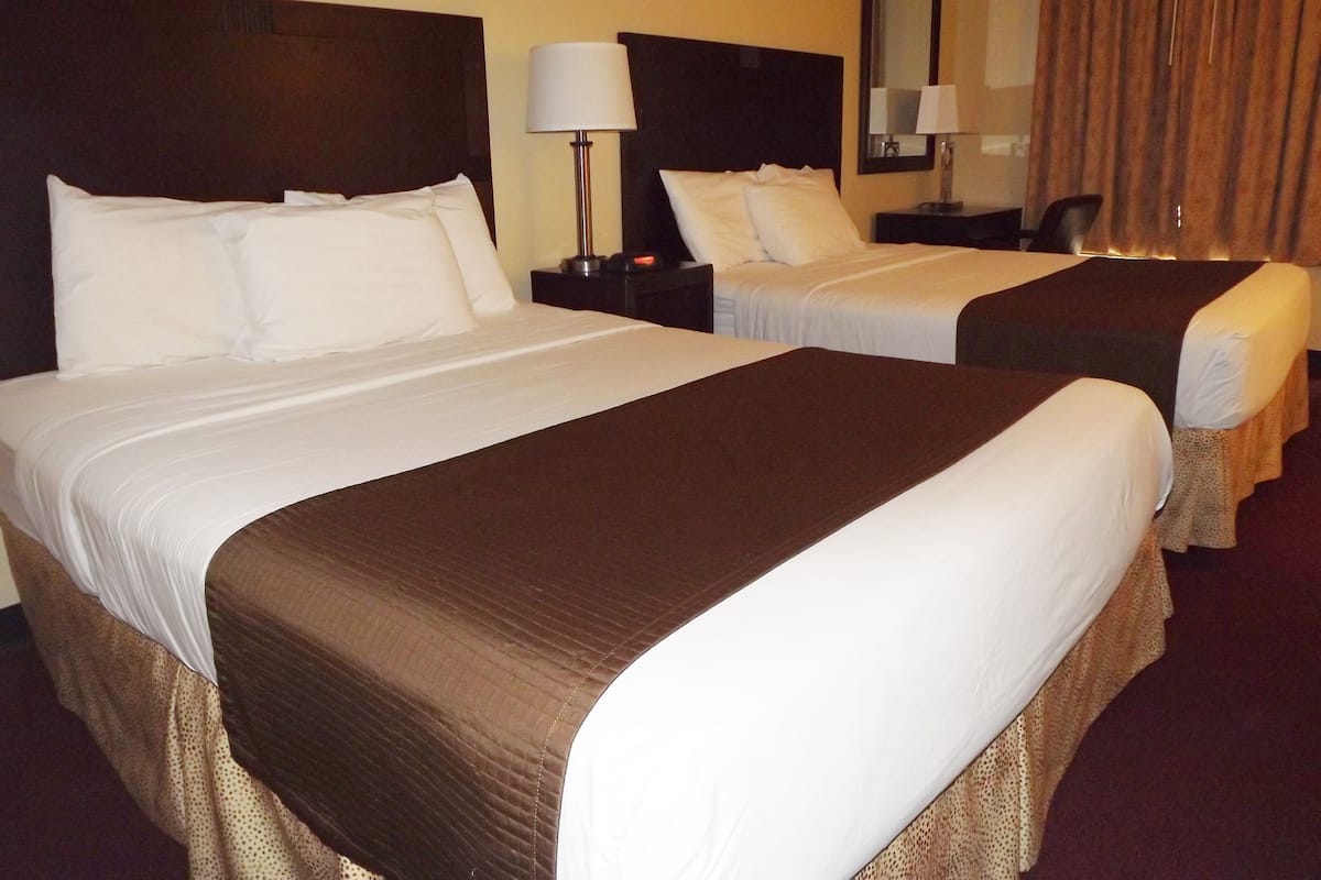 A hotel room with two beds, each with white linens and brown headboards. There is a nightstand with a lamp between the beds.