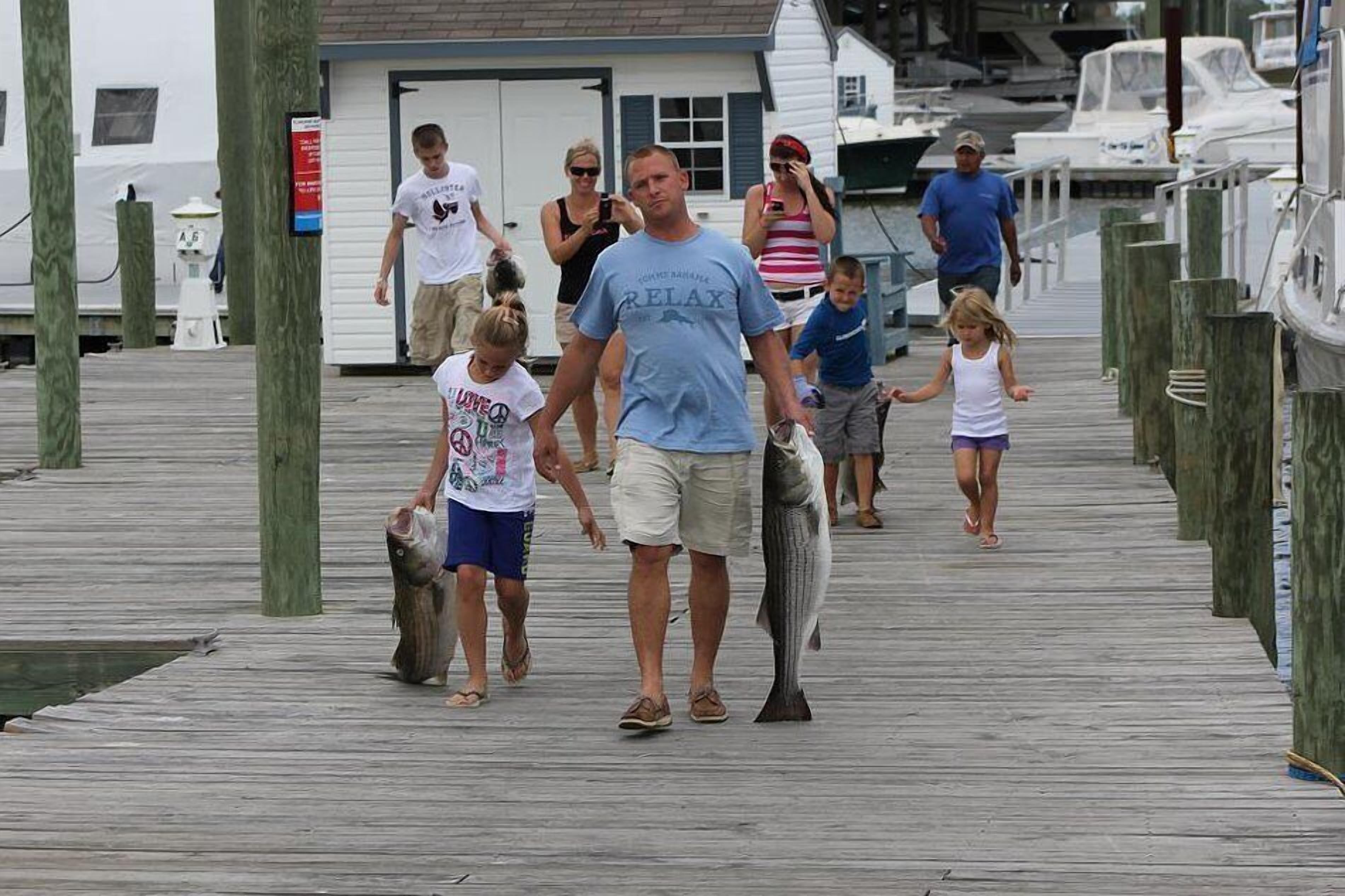 A wooden pier with people walking, boats docked nearby, and a white building in the background.