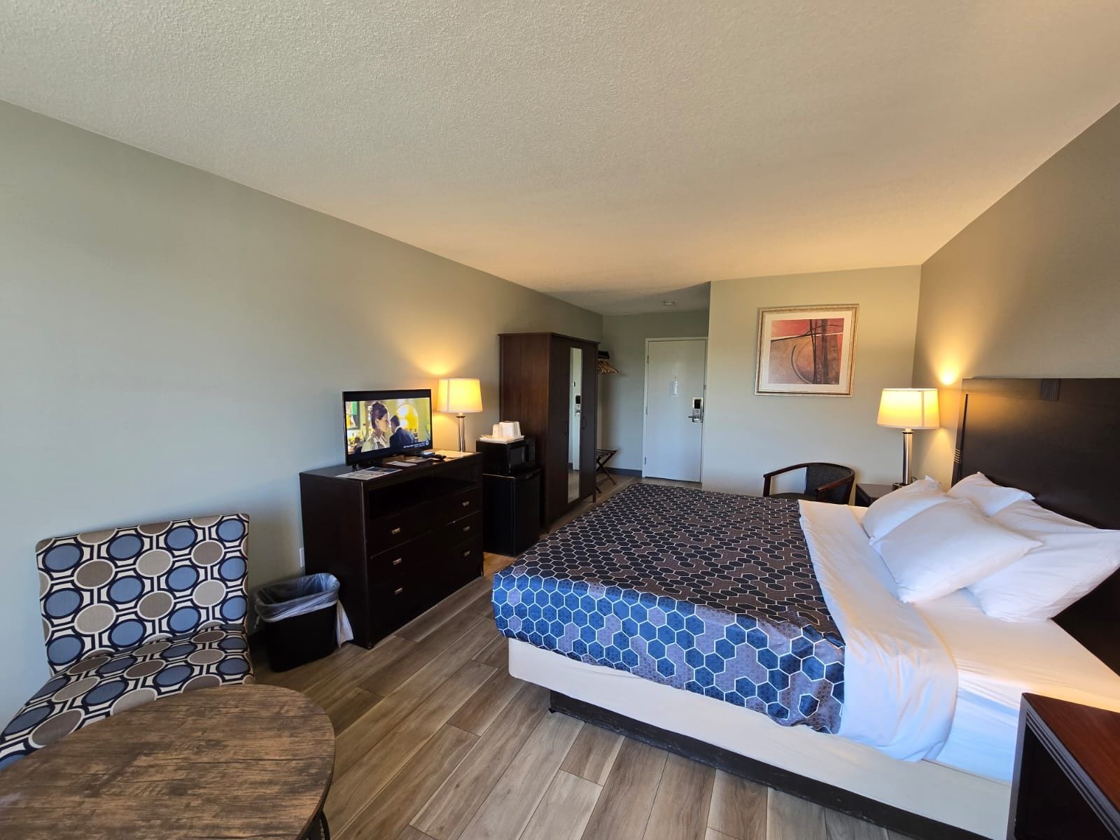 A hotel room with a bed, a desk with a television, a chair, and a door leading to another room.