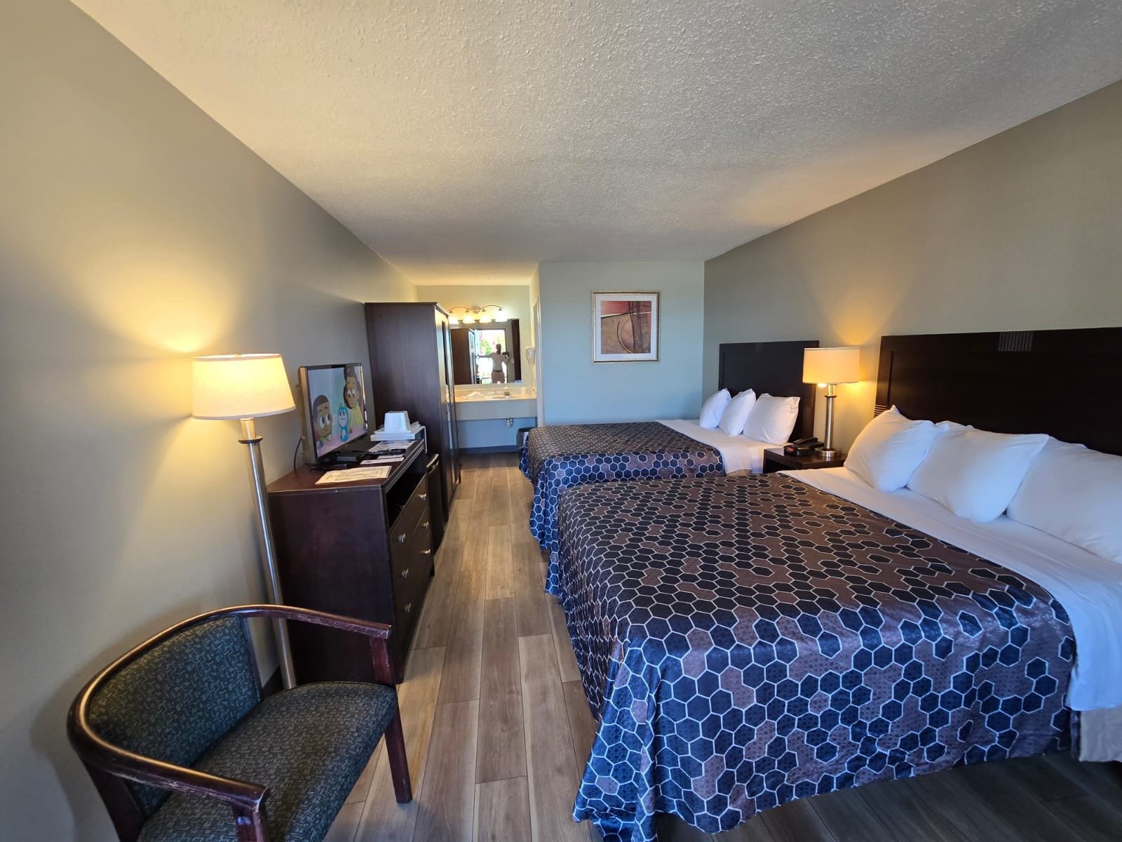 A hotel room with two beds, a desk with a chair, a mirror, and a lamp.