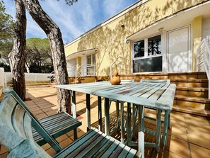 An outdoor patio area with a wooden table and benches, a large tree, and a yellow building with a window and a door. - Brisa CostaBravaSi - Beach, A/C, Parking (Platja d'Aro)