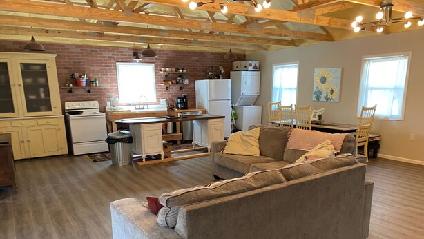 A spacious living area with a grey sofa, wooden flooring, a kitchen with a brick wall, and a dining area with a wooden table and chairs. - Edina Missouri New Cabin in Rural Area (Edina)