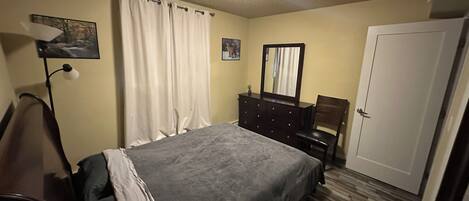Un dormitorio con cama, cómoda, silla, escritorio, lámpara y puerta.