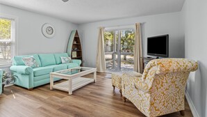 Ein Wohnzimmer mit einem türkisfarbenen Sofa, einem gemusterten Sessel, einem Flachbildfernseher, einem Deckenventilator und einem großen Fenster mit Vorhängen.