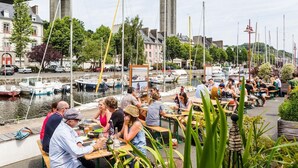 Un espace de restauration au bord de la rivière, avec des tables et des chaises, des bateaux amarrés le long de l’eau et un pont en arrière-plan.