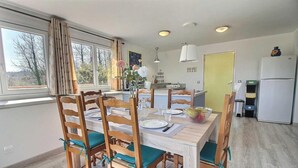 Un comedor con una mesa y sillas de madera, una cocina al fondo y vistas a árboles a través de la ventana.