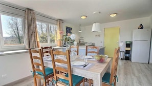 A dining area with a wooden table and chairs, a window with curtains, a kitchen with a refrigerator, and a yellow door.