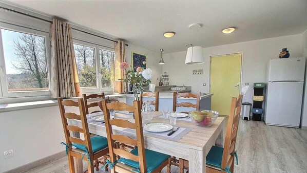 A dining area with a wooden table and chairs, a window with curtains, a kitchen with a refrigerator, and a yellow door. - Le Nid D'hirondelles (BOURBRIAC)