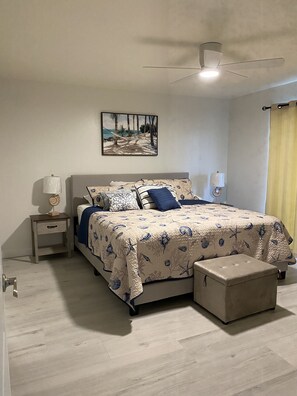 Un dormitorio con cama, mesitas de noche, ventilador de techo, un cuadro y una ventana con cortinas.