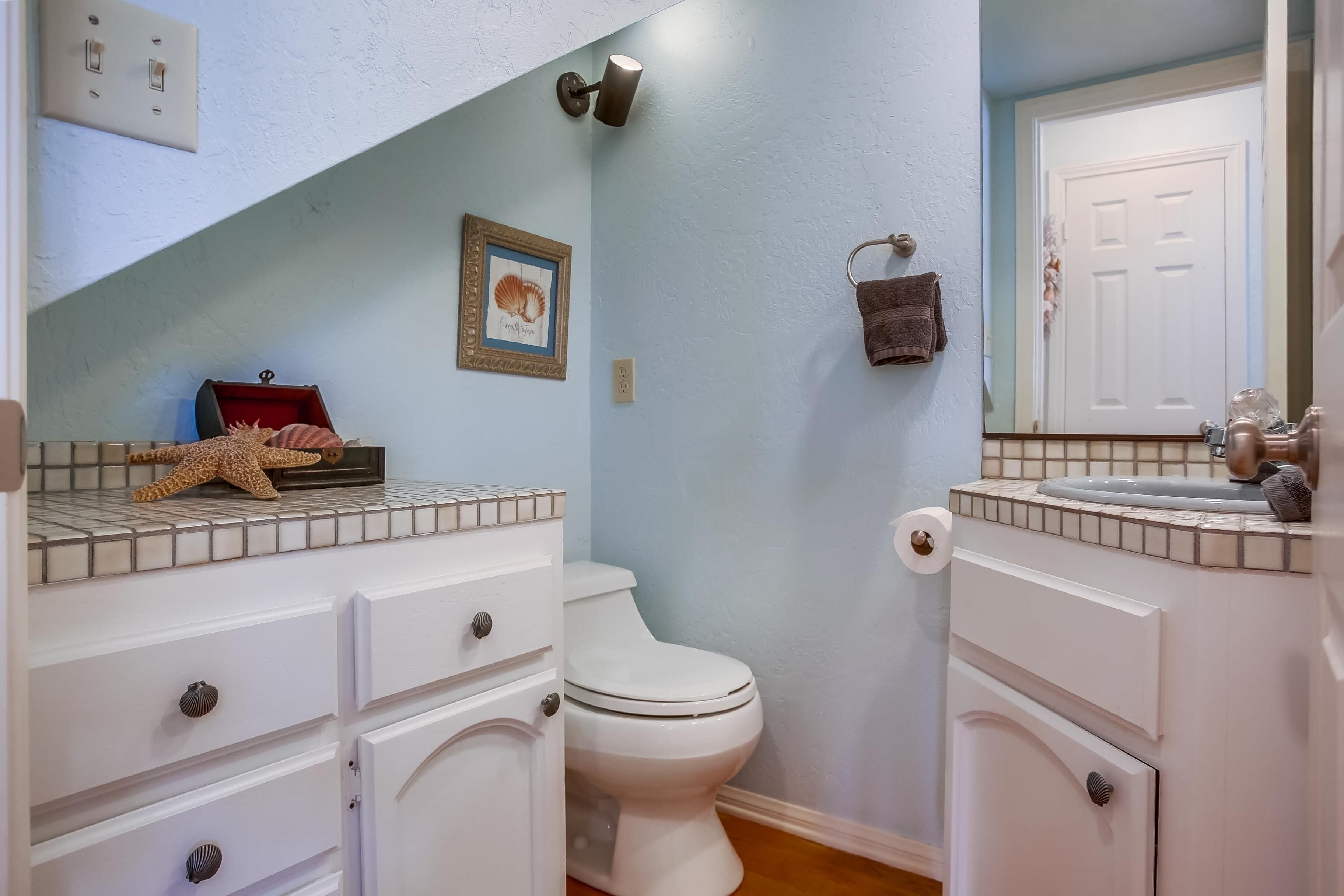 Casa de banho com sanita branca, lavatório branco e espelho sobre o lavatório. Há um quadro emoldurado na parede e um porta-toalhas com uma toalha pendurada.
