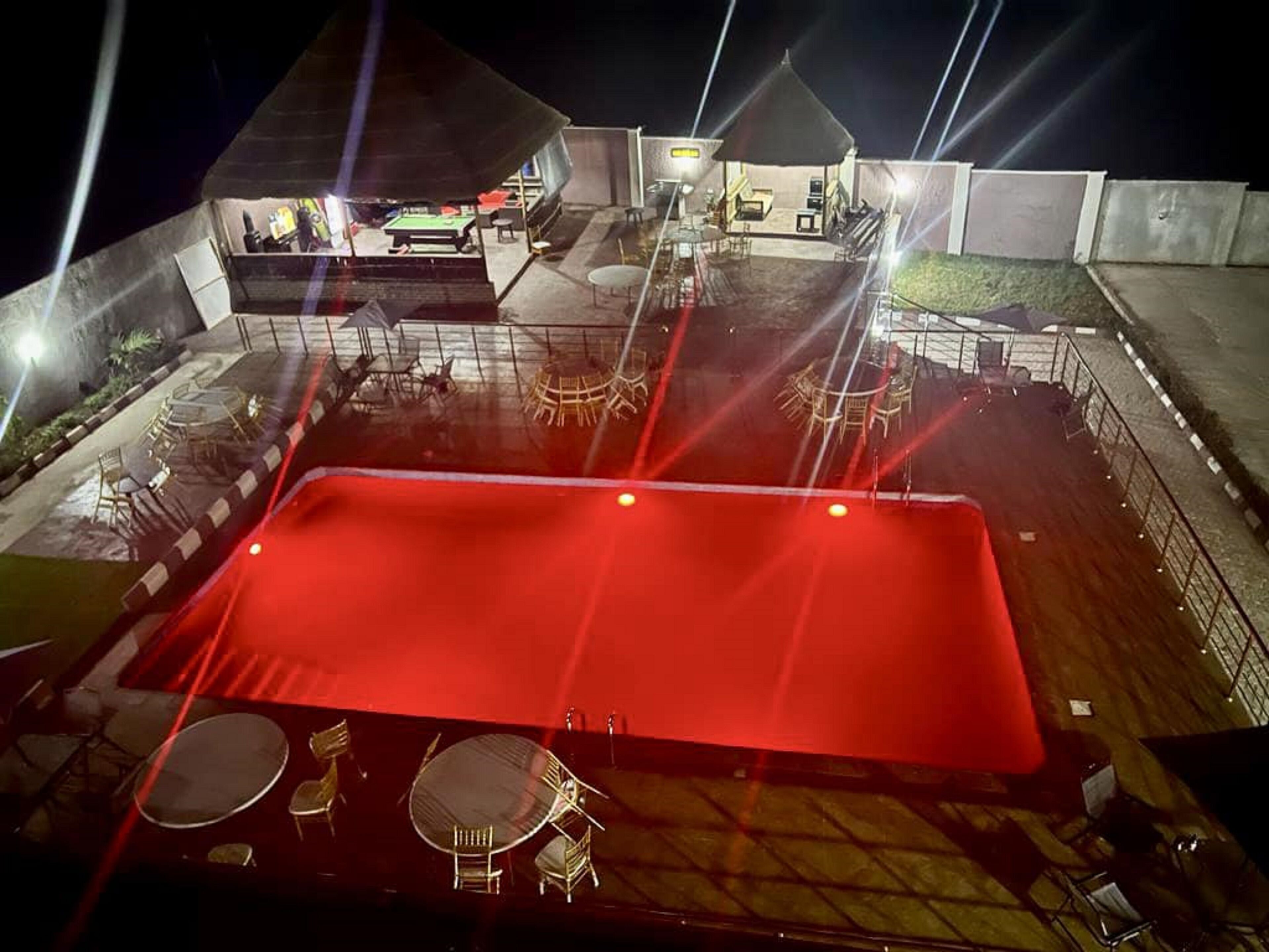 Un área de piscina al aire libre con una cubierta roja, rodeada de mesas y sillas, y un edificio pequeño al fondo.