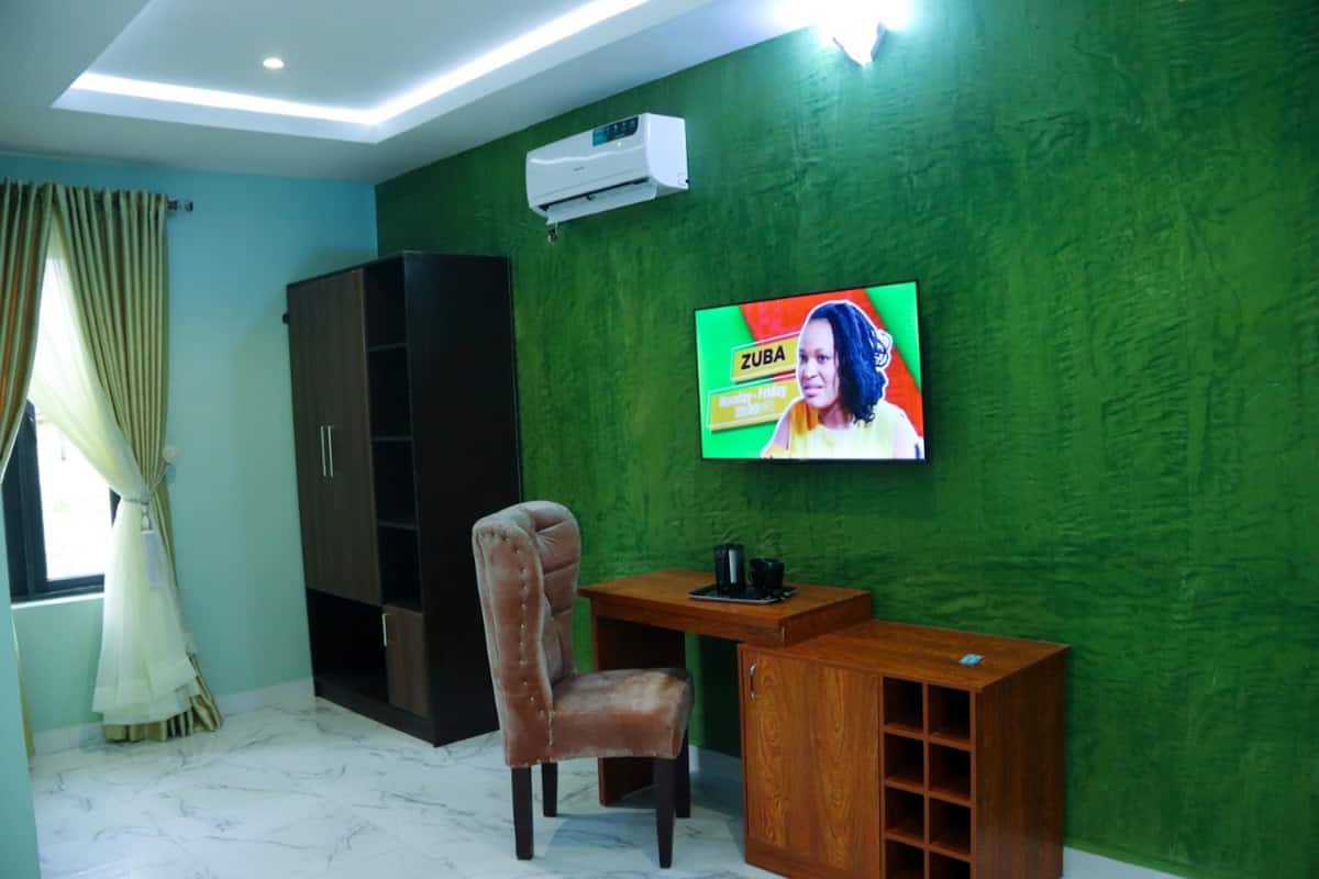 Quarto com parede verde texturizada, televisão fixada na parede, escrivaninha de madeira, poltrona de couro marrom e guarda-roupa de madeira escura.