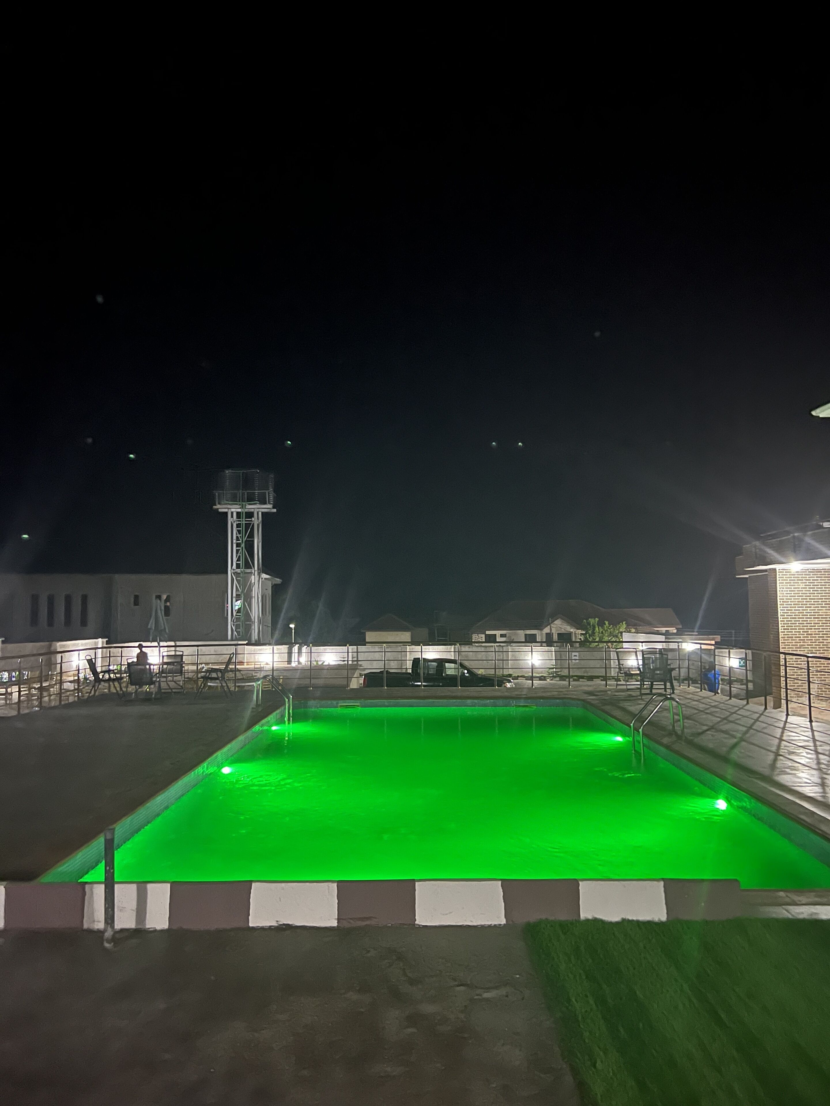Una piscina con elemento d'acqua illuminato di verde, circondata da un'area recintata con sedie e tavoli, e un'alta torre d'acqua sullo sfondo.