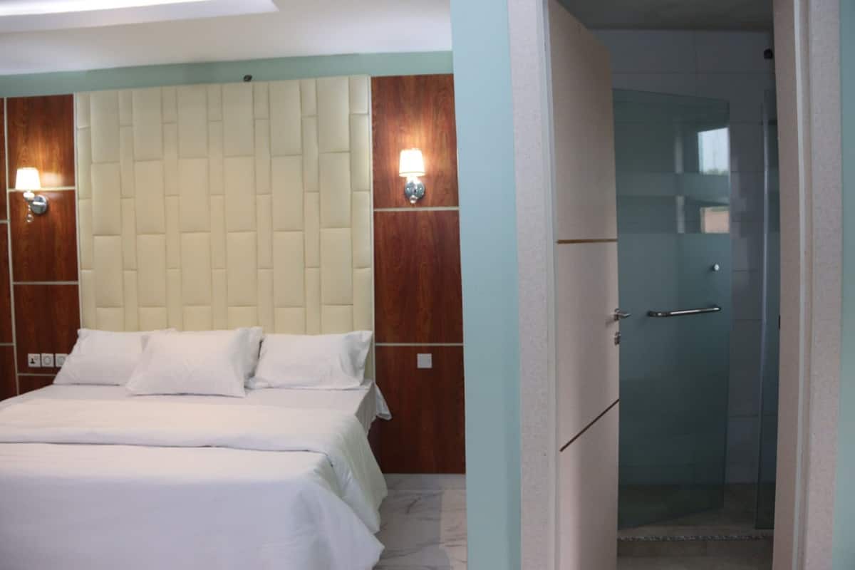 Quarto de hotel com cama, cabeceira de madeira, duas luzes fixadas na parede e porta de vidro para o chuveiro.