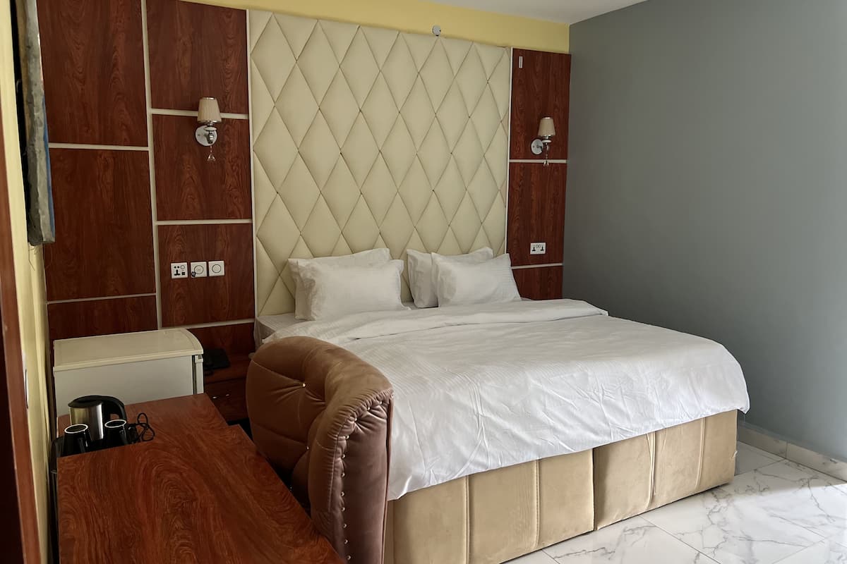 Quarto com cabeceira estofada, armário de madeira, mesa de cabeceira com chaleira e cama com roupa de cama branca.