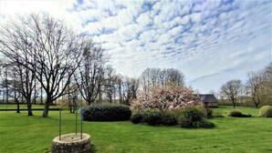A well-maintained grassy area with a stone monument, flowering bushes, and leafless trees under a partly cloudy sky.