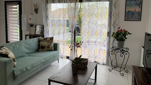 A living room with a sofa, a coffee table, and a potted plant. There is a window with curtains and a framed picture on the wall.