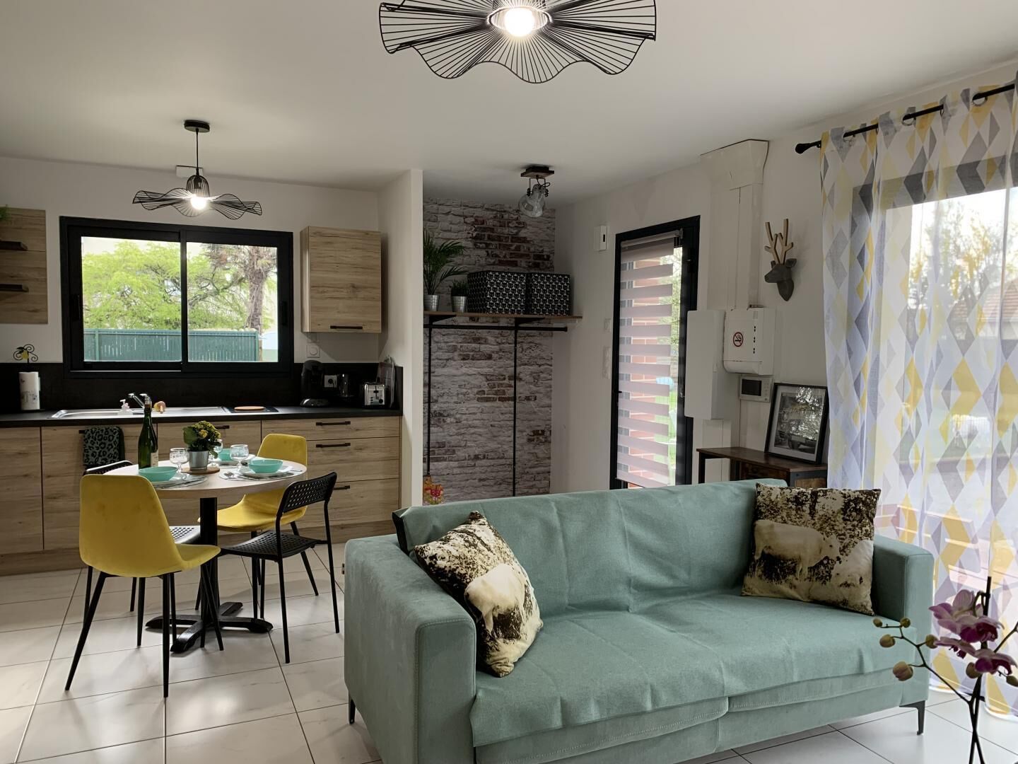 A modern kitchen with a dining area, a sofa, and a view of greenery outside.