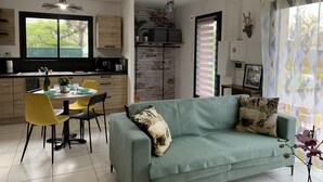 A modern kitchen with a dining area, a sofa, and a view of greenery outside.