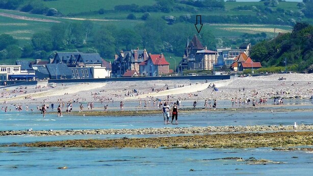 Eine Küstenstadt mit Sandstrand, Häusern mit roten Dächern und einer hügeligen, grünen Landschaft im Hintergrund.