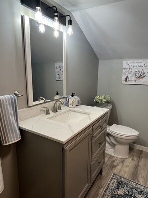 A modern bathroom with a large mirror, two light fixtures, a sink, a toilet, and a decorative sign on the wall. - Calla Lily #2 - Oneonta Apartment close to downtown - 1 of 2 units (Oneonta)
