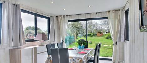 Un coin repas avec une table dressée pour deux personnes, une vue sur le jardin et un espace cuisine avec une fenêtre.