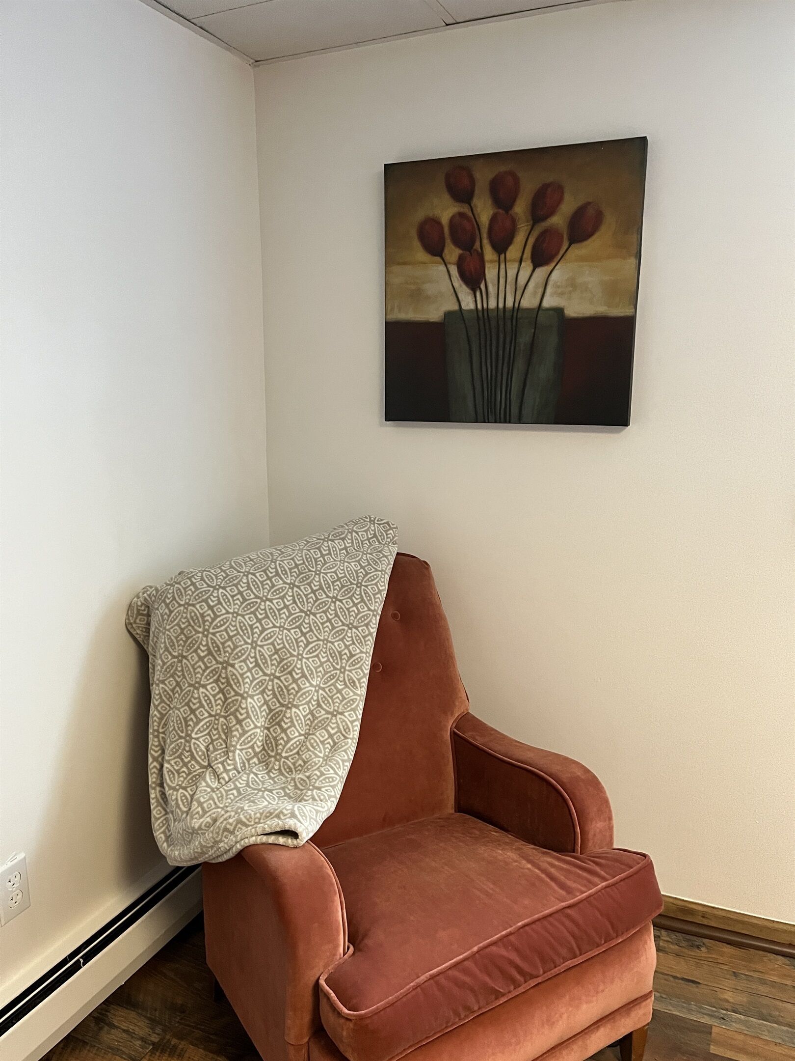 A corner room with a red velvet armchair, a patterned throw, and a painting of red flowers on the wall.