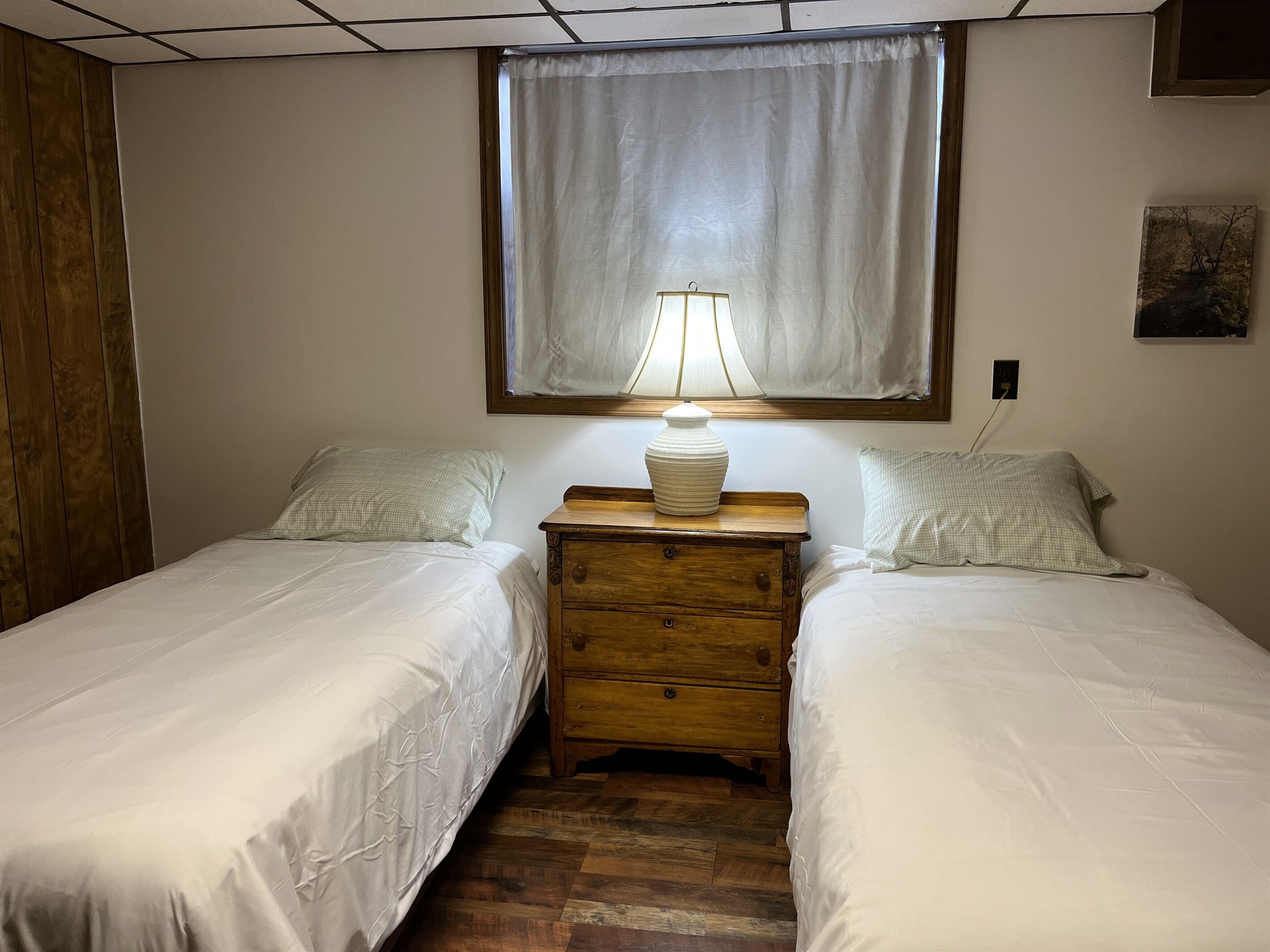A bedroom with two single beds, a wooden nightstand with a lamp, a window with a sheer curtain, and a small painting on the wall.