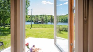 En vy från insidan av ett rum med en träveranda, med utsikt över en gräsyta med en liten byggnad och en sjö.