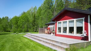 En röd stuga med en träveranda, omgiven av ett frodigt, grönt landskap och höga träd.