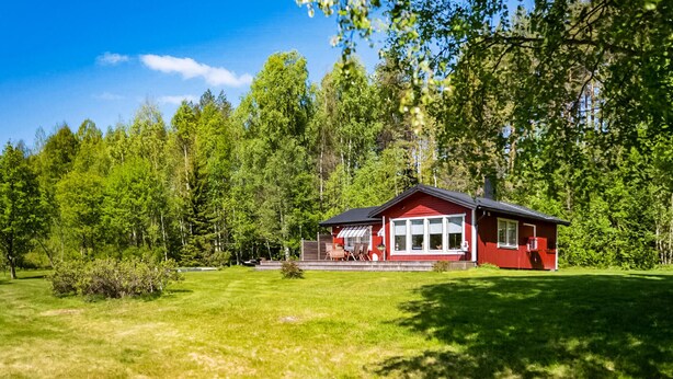 En röd stuga med en veranda, omgiven av en gräsklädd trädgård och en tät skog.