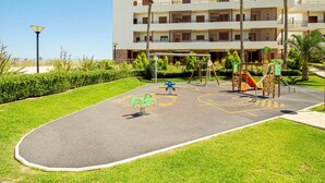 Un parque infantil con varios juegos, rodeado de un césped cuidado y palmeras, frente a un edificio de varios pisos.