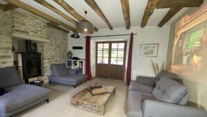A cozy living room with a stone wall, wooden beams, a fireplace, and a large window with curtains. - Longère *** with large heated swimming pool, sports room, billiards table, fibreglass in the countryside (La Roche-l'Abeille)