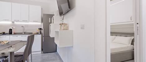 Een moderne keuken met witte kasten, een eethoek met een tafel en stoelen, en een slaapkamer met een bed en een televisie aan de muur.