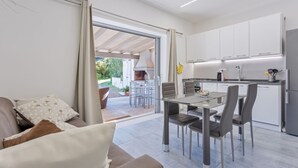Eine moderne Küche mit weißen Schränken, einem Essbereich mit einem Glastisch und einer Glastür, die auf eine Terrasse im Freien führt.