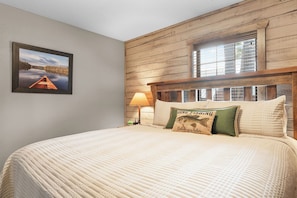 Une chambre à coucher avec un grand lit, une tête de lit en bois, une fenêtre avec des stores, un tableau encadré au mur et une table de chevet avec une lampe.