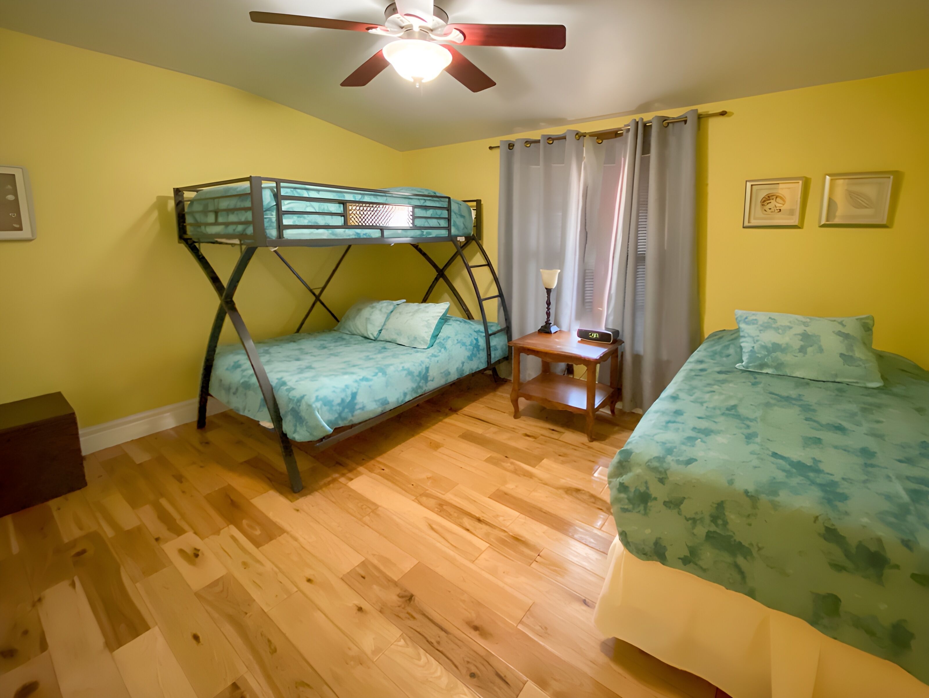 A room with a bunk bed, a single bed, a wooden nightstand, and a ceiling fan.