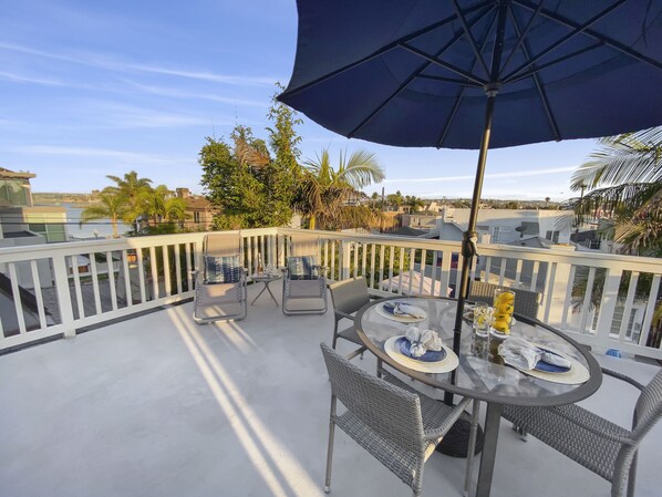 A rooftop patio with a round glass table, two chairs, a large umbrella, and a view of the cityscape.