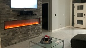 A modern living room with a stone wall, a flat-screen TV mounted above a fireplace, and a glass coffee table in the foreground. - Modern comfort, small town charm. Room for 
all to visit, rest retreat or create (Center Point)