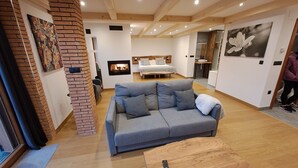 Ein modernes Wohnzimmer mit grauem Sofa, Kamin, Bett mit Holz Kopfteil und Küchenbereich im Hintergrund.