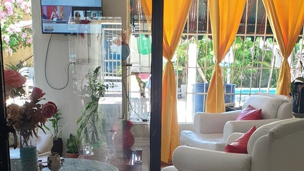 Uma sala de estar com um sofá branco, uma mesa redonda com toalha de mesa estampada e uma televisão na parede.