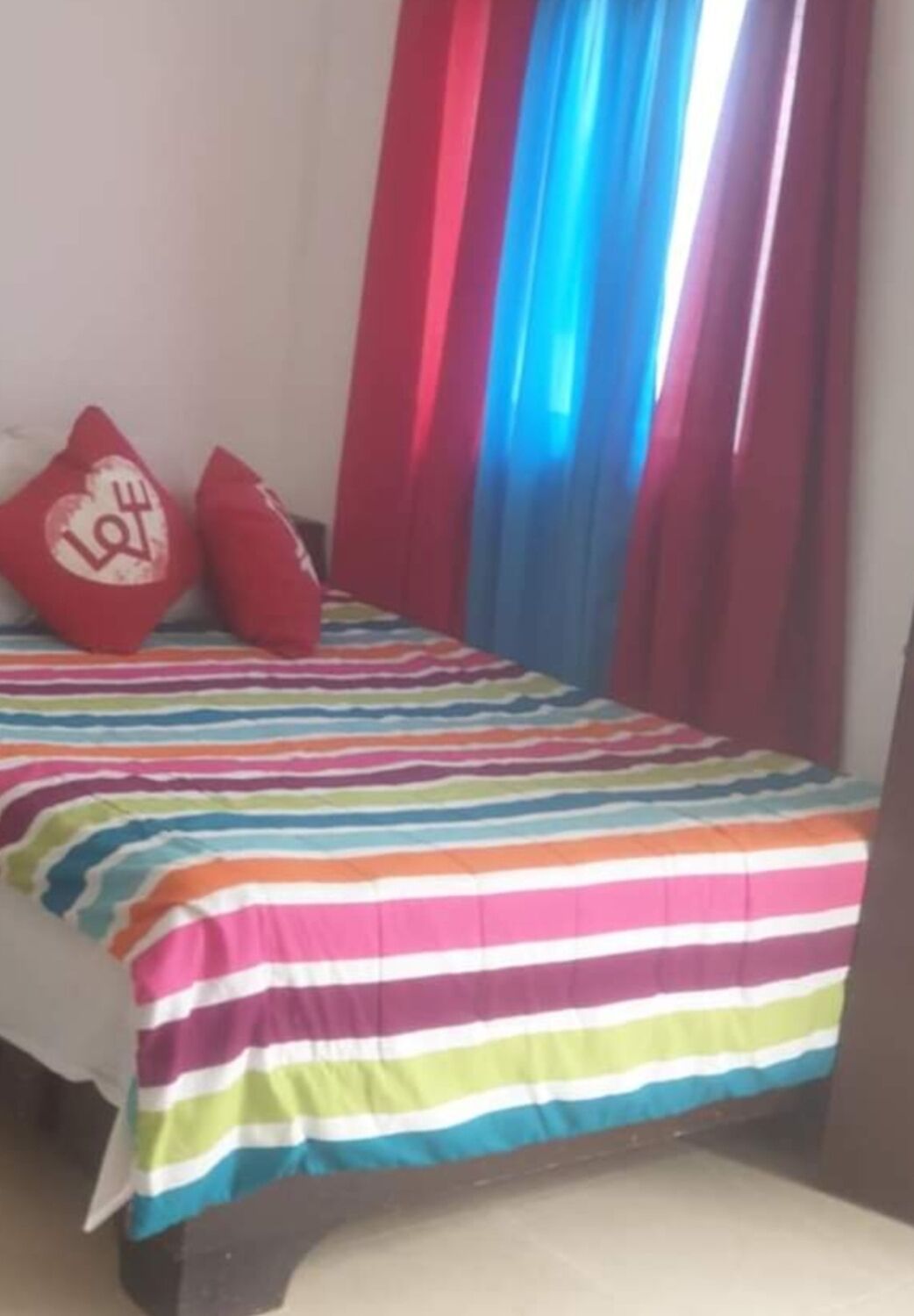 A bed with a multicolored striped bedspread, two red pillows with a heart design, and red curtains with a blue and white pattern.