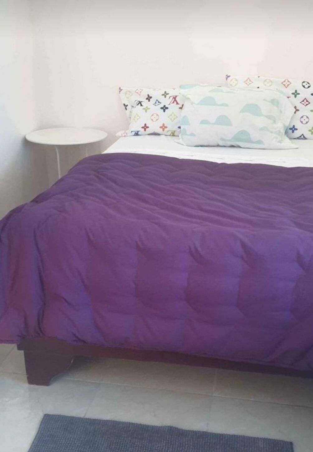 A bed with a purple quilt, a white pillow with a pattern, and a small round table beside the bed.