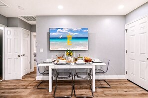 Un coin repas avec une table dressée pour quatre personnes, une vue dégagée sur l’océan et une peinture représentant un voilier au mur, ainsi qu’un design moderne avec des murs blancs et un plancher en bois.