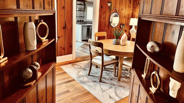 A wooden-paneled room with a dining table, chairs, and a kitchen in the background.