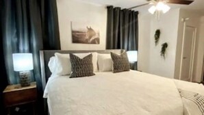 A bedroom with a large bed, two bedside tables with lamps, a ceiling fan, and a wall-mounted artwork.