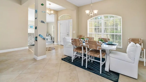 A dining area with a white table and chairs, a chandelier, and a large window with blinds.