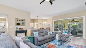 A spacious living room with a grey sofa, a patterned armchair, and a colorful rug. There is a dining area with a table and chairs, and a large sliding glass door leading to an outdoor area.