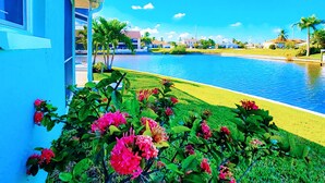 A poolside view with a clear blue sky, a building with a blue exterior, and pink flowers in the foreground. - 30% Disc-2 Kg Mstrs, Golf, Fishing, BBQ, Pool, Spa (Cape Coral)