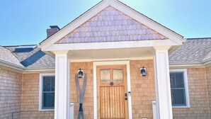 A house with a front porch, a wooden door, and two wall-mounted lights. - Secluded, Private Vacation Beach Home on Chappaquiddick Island, Walk To Beach. (Edgartown)